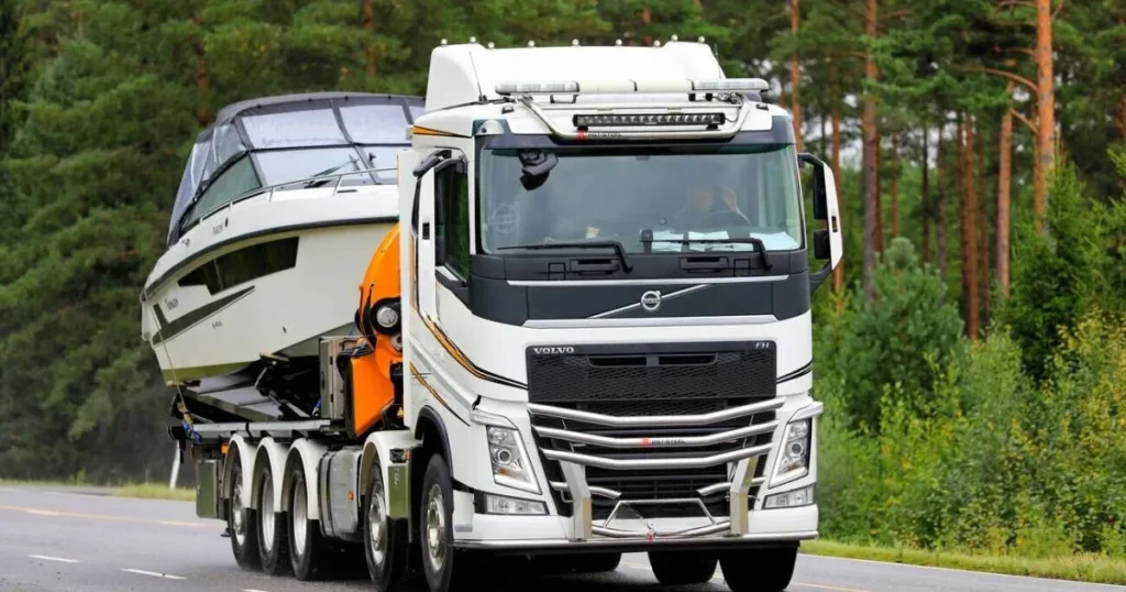 "Volvo FH truck performing Boat Transport in the UK, carrying a large boat on a highway through a forested area"